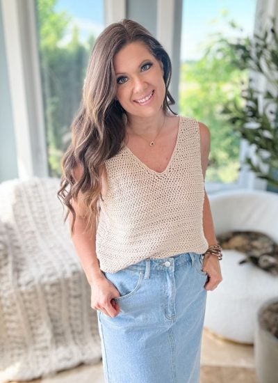 A woman stands indoors, wearing a beige knit top reminiscent of the Crochet Ruby-V Tank and a blue denim skirt. Her long brown hair cascades as she smiles warmly. In the background, a cream-colored chair and lush green foliage add to the serene scene.