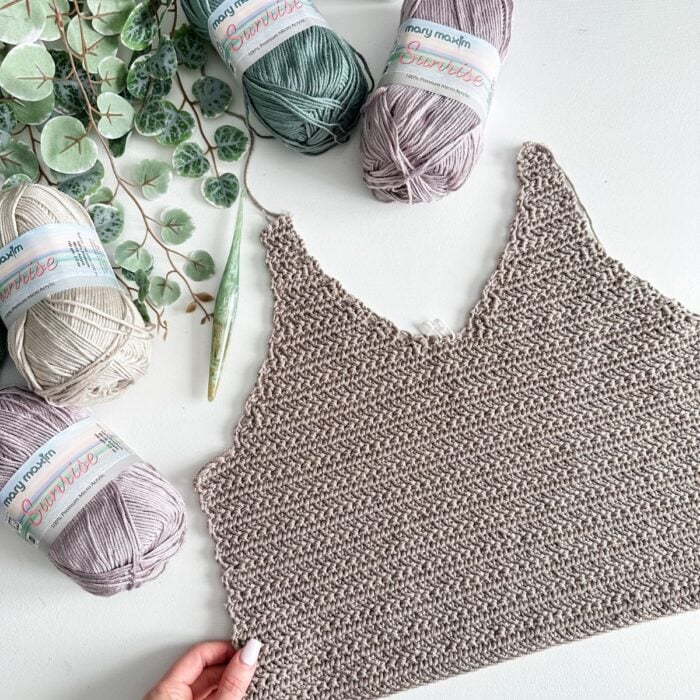 A hand holding a knitted piece lies near four skeins of yarn—one green and three shades of beige—on a white surface decorated with green leaves, ready to Crochet the Ruby-V Tank.