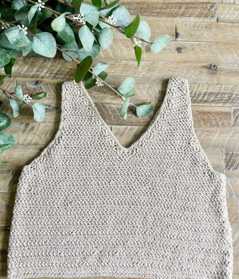 A beige, hand-knitted Crochet the Ruby-V Tank with a V-neck is laid flat on a wooden surface, next to a leafy green plant branch.
