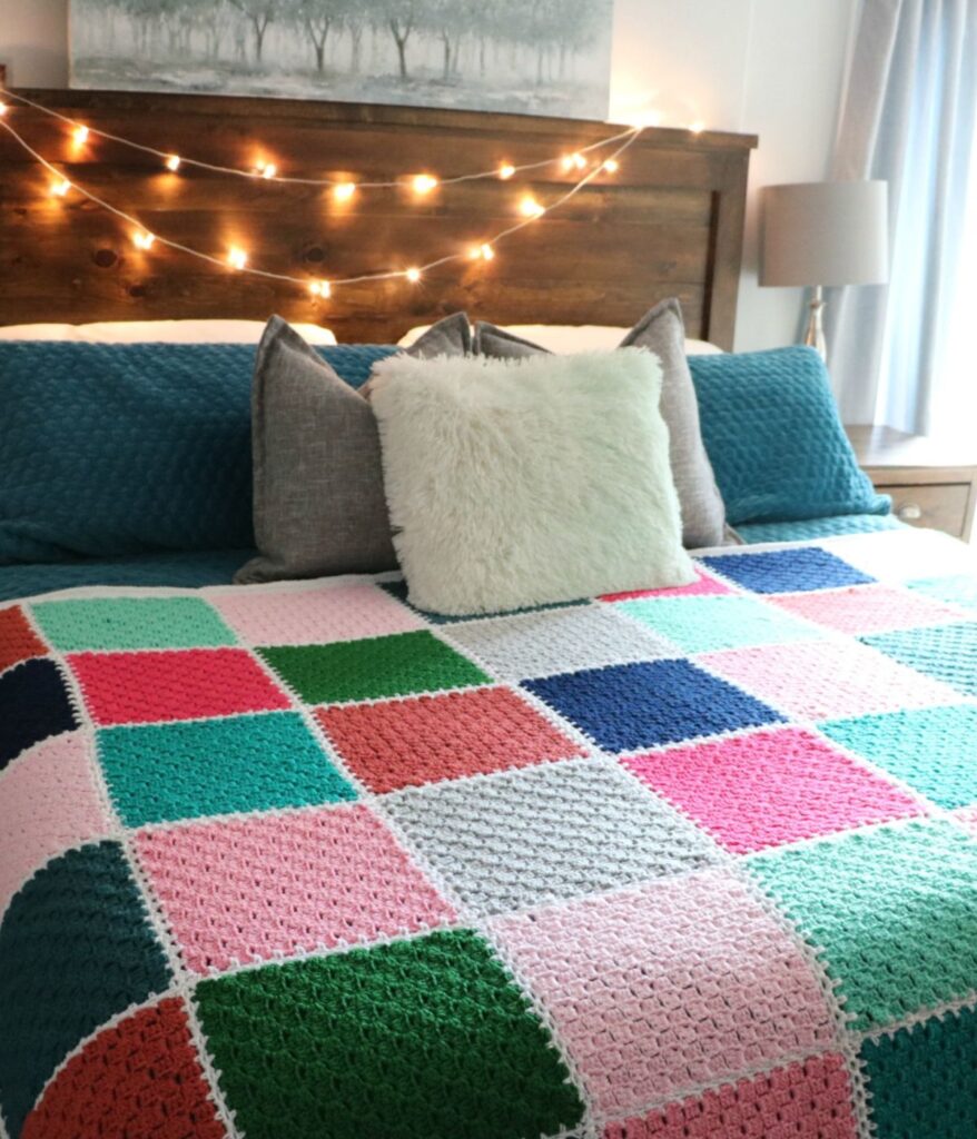 A bed with a Modern Patchwork Crochet Throw, colorful pillows, a wooden headboard, string lights, and a bedside lamp.