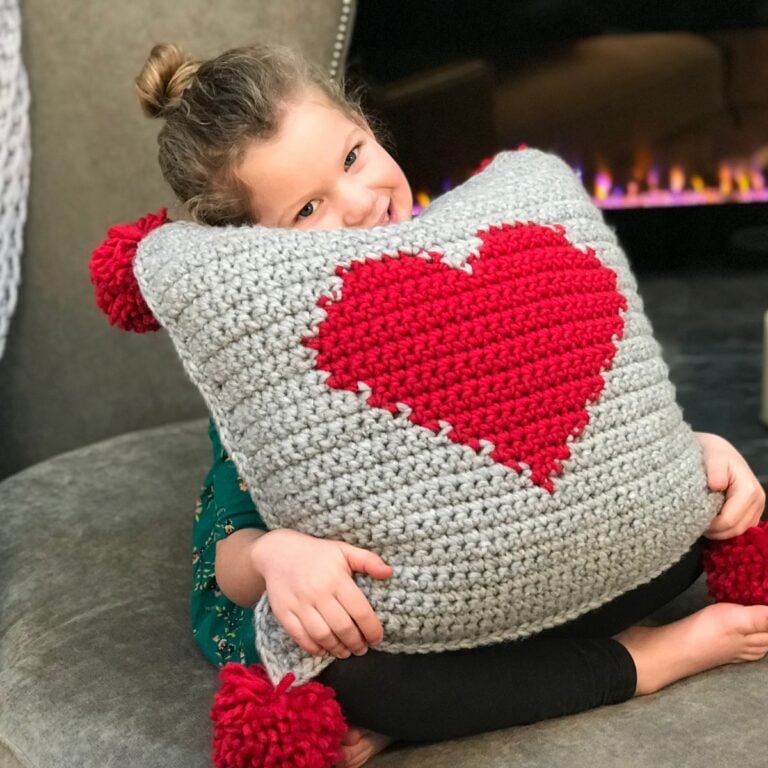 A little girl holding a crocheted heart pillow in front of a fireplace.