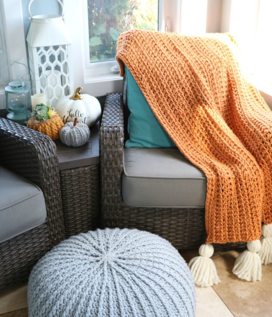 A wicker chair with a gray cushion and blue pillow is draped with a cozy orange Crochet Chunky Blanket. Nearby are knit pumpkins, a white lantern, and a light gray knit pouf.