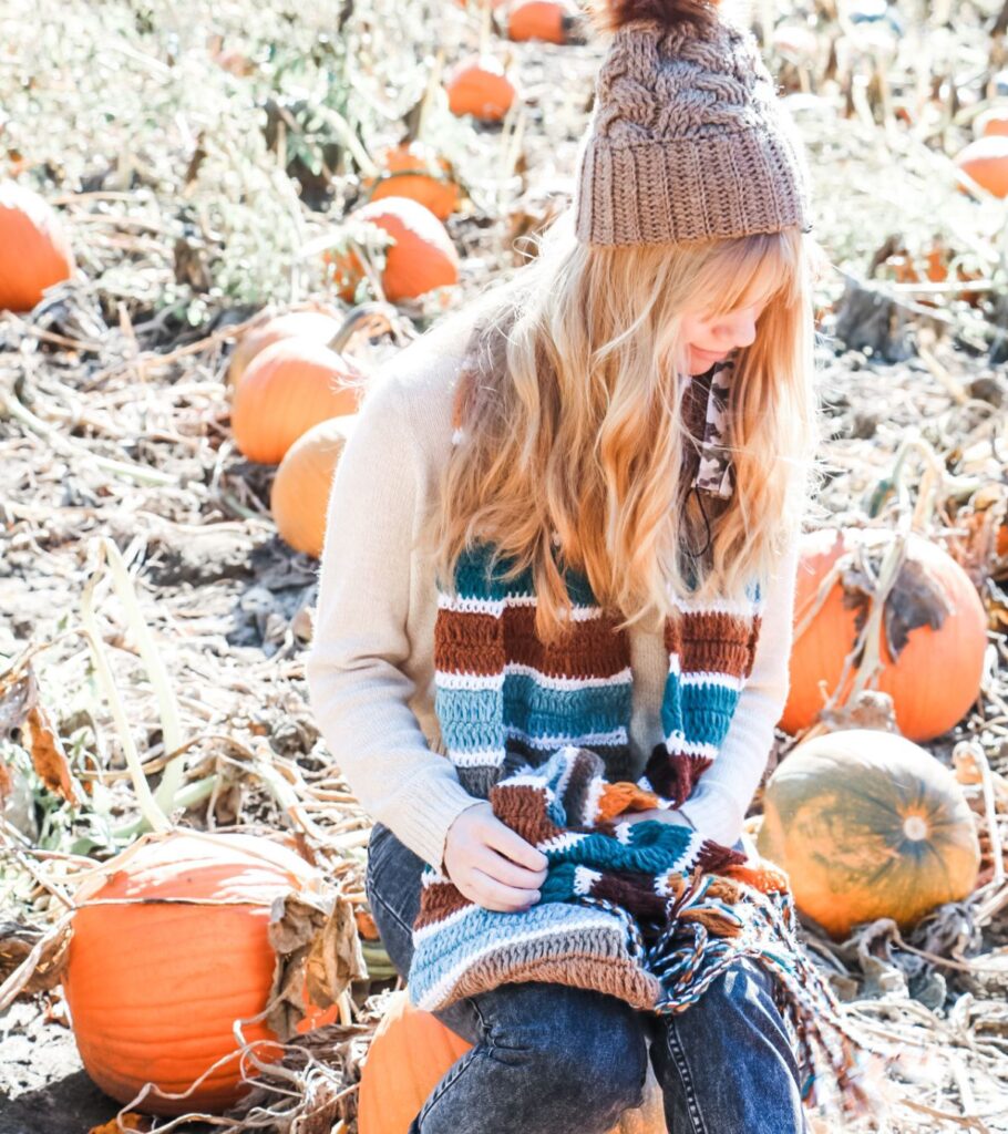 A girl sitting in a cozy pumpkin patch, wrapped in a crochet blanket scarf.