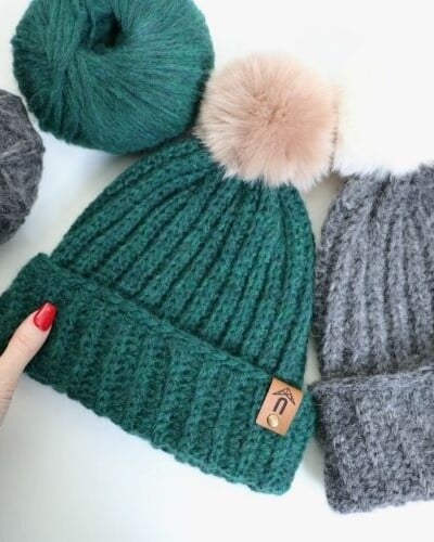 A person is crocheting a beanie hat with yarn and pom poms.