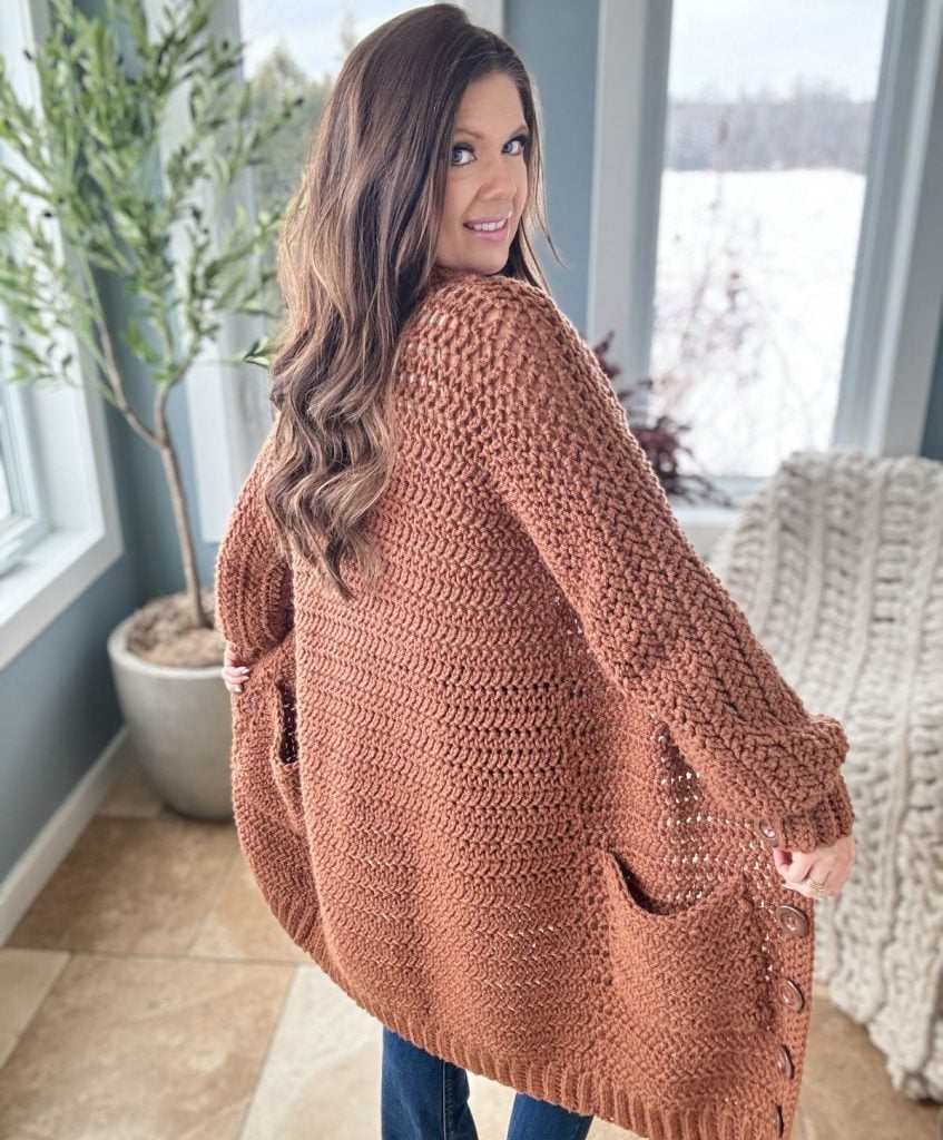 Woman with long hair wearing a loose, brown knit cardigan over a crochet crop top pattern, standing in a room with large windows and a plant. She looks over her shoulder, facing the camera.