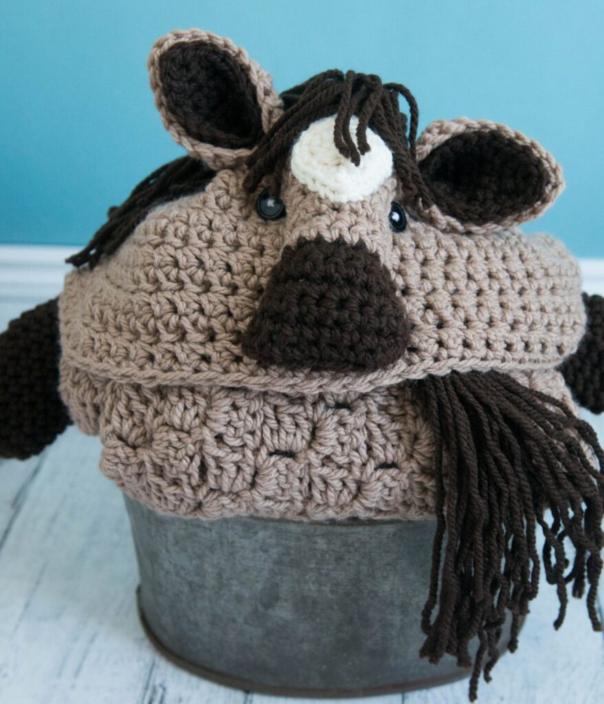 A crocheted hat designed to look like a brown and tan animal with ears, a snout, a white horn, and yarn for hair sits atop a gray metal container—perfect for fans of whimsical accessories or pairing with a cozy crochet horse blanket.