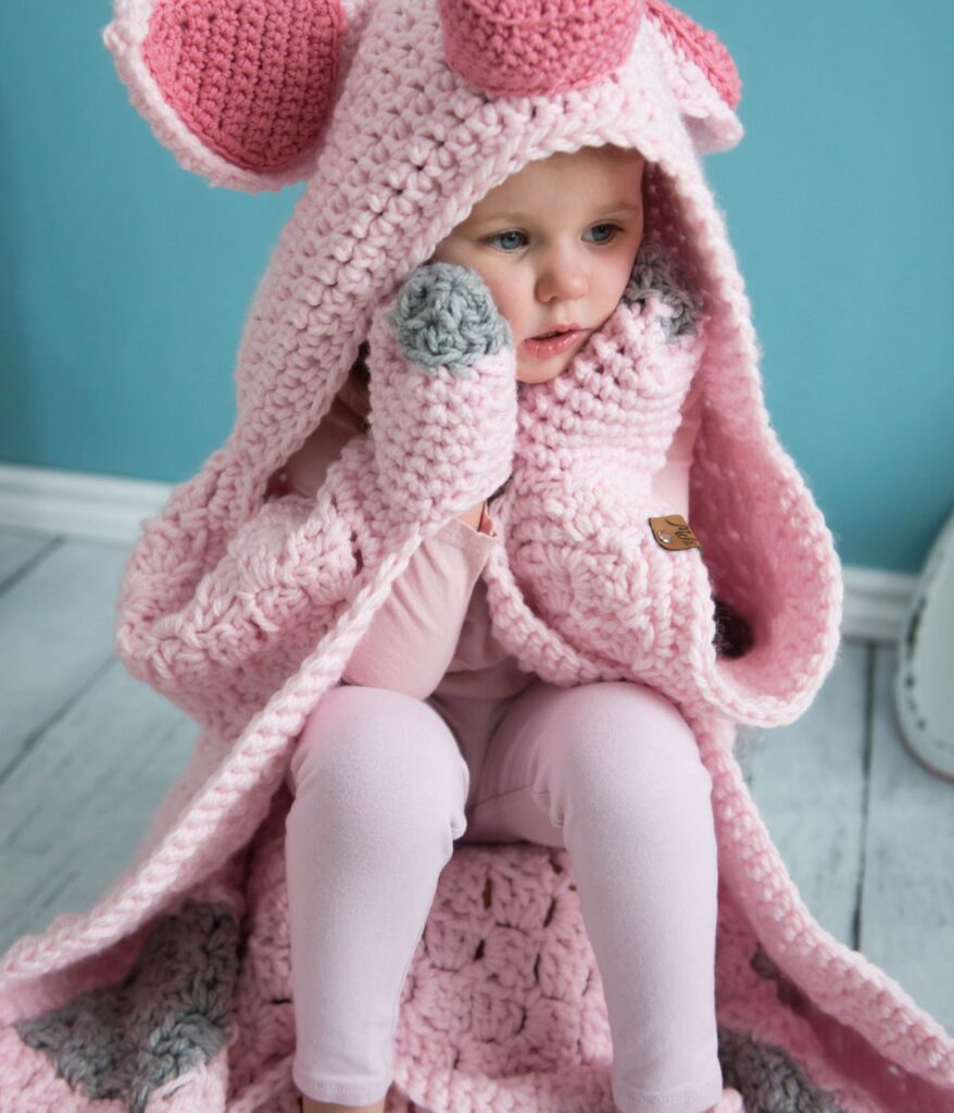 A young child sits on the floor wearing a pink Crochet Pig Blanket with animal ears and paws, looking to the side.