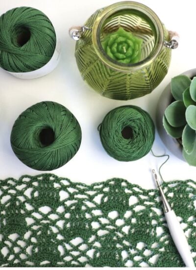 Green yarn spools, a crochet hook with in-progress sleeveless cardigan pattern, and a succulent next to a translucent green candle holder on a white surface.