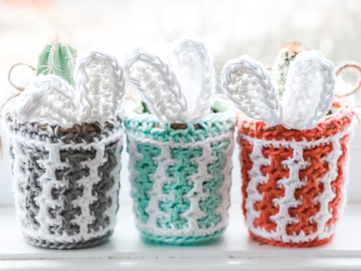 Three Easter-themed crocheted cacti with white flowers in multicolored pots displayed in front of a window.