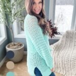Woman in a teal crochet cardigan and jeans, smiling in a room with a snowy view outside the window.