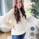 Woman in a white crochet sweater and distressed jeans standing in a room with a window view, hand on hip, smiling at the camera.