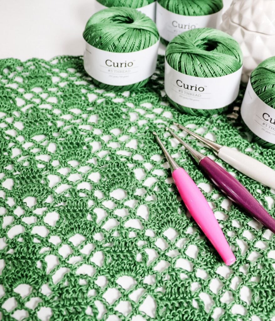 Green crocheted sleeveless cardigan pattern with a crochet hook, scissors, and three balls of green yarn labeled "curio" on a white background.