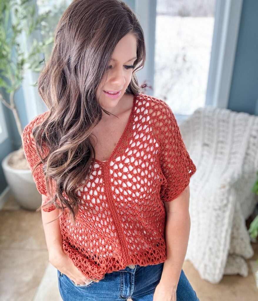 A woman with long brown hair, wearing a Rusty Road crop top and jeans, looks down while standing indoors near a doorway.