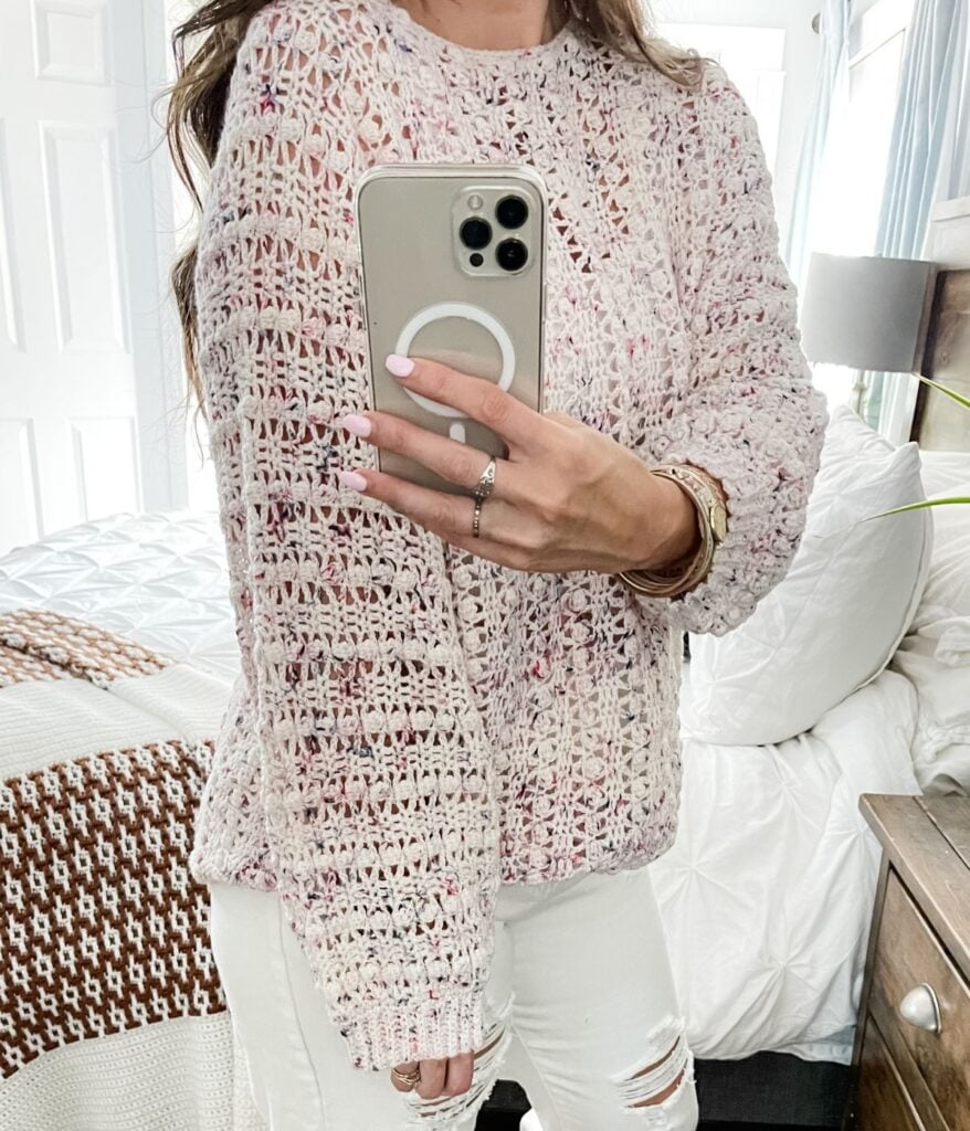 Woman holding a smartphone wearing a chunky, multicolored Gum Drop Pullover and white ripped jeans.