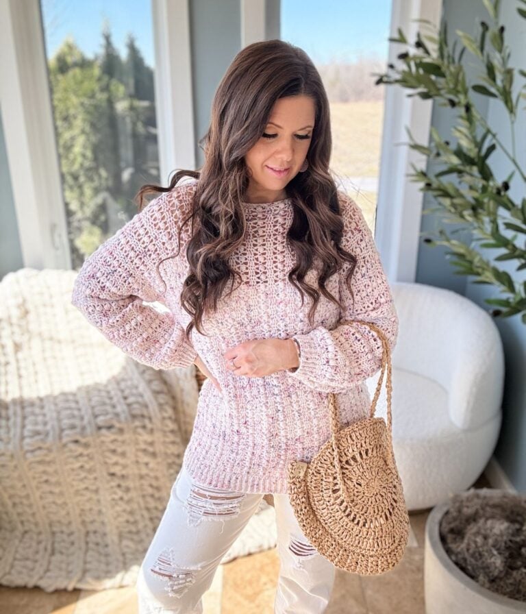 A woman in a Gum Drop Pullover and white jeans carrying a straw handbag standing by a window.