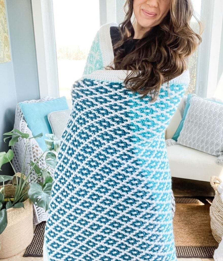A woman stands indoors wrapped in a thick, blue and white patterned Caribbean Coast Afghan, with plants and cushions visible in the background.