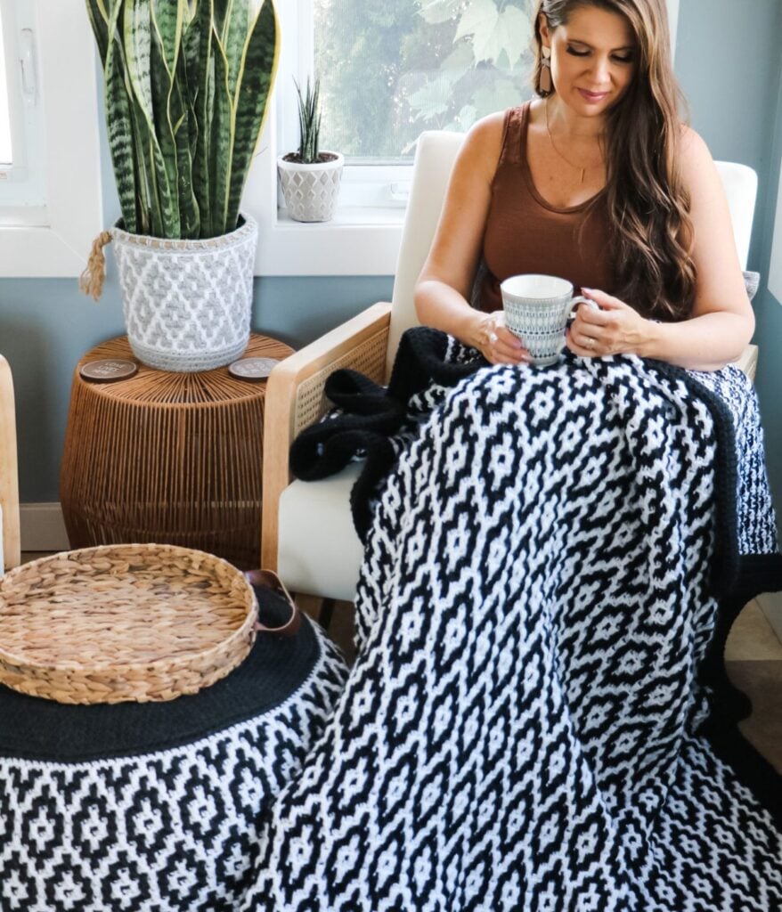 A woman sits in a modern chair, wrapped in a Granite Ridge Mosaic Blanket, holding a mug. A potted plant and wicker table are nearby in the bright room.