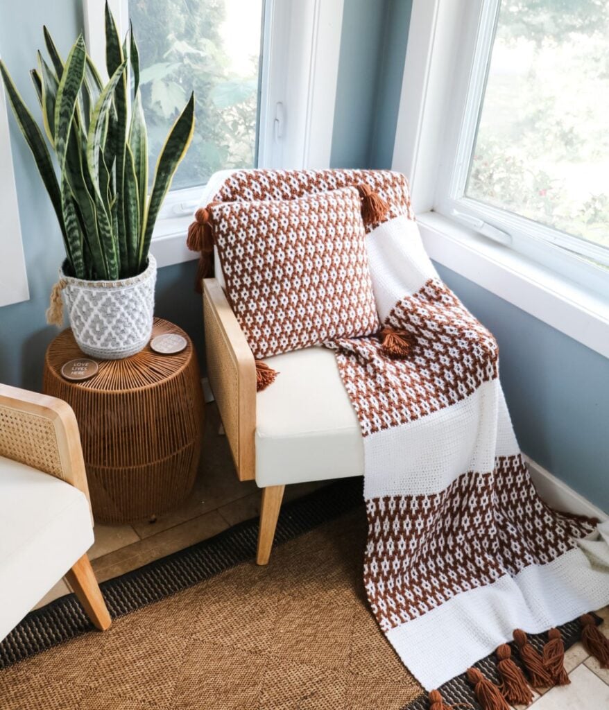 A Desert Dunes Mosaic Blanket and matching pillow add a patterned touch to a cream armchair, set beside a potted snake plant on a wicker side table by the window.