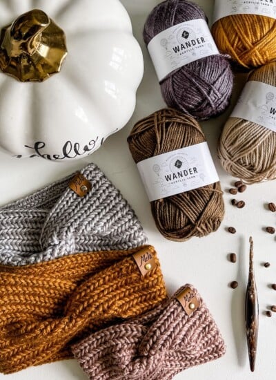 A Harvest Twist Ear Warmer sits on a table, surrounded by a pumpkin, yarn, and knitting needles.