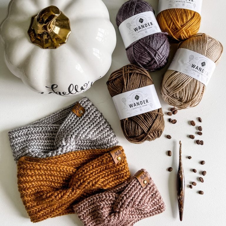 A Harvest Twist Ear Warmer sits on a table, surrounded by a pumpkin, yarn, and knitting needles.
