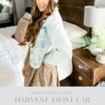 Woman wearing the Harvest Twist Ear Warmer, a light denim jacket, and a printed dress stands in a bedroom holding a basket. Text overlay reads "Harvest Twist Ear Warmer.