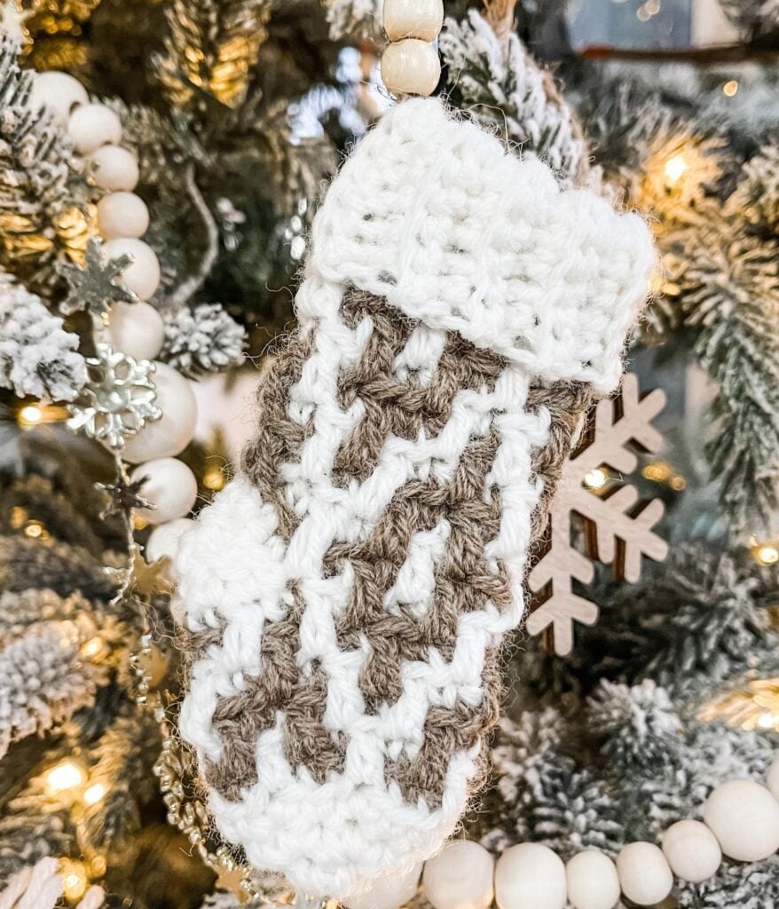 A crocheted mitten ornament in white and beige hangs on a Christmas tree alongside a Mini Mosaic Stocking, surrounded by beads, snowflakes, and twinkling lights.