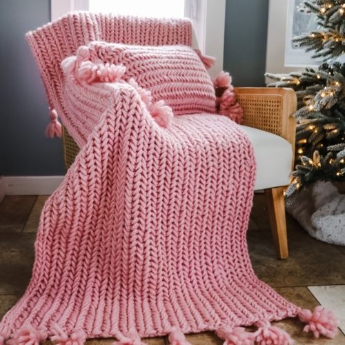 A pink knitted blanket with tassels in front of a christmas tree, perfect for cozy social media posts.