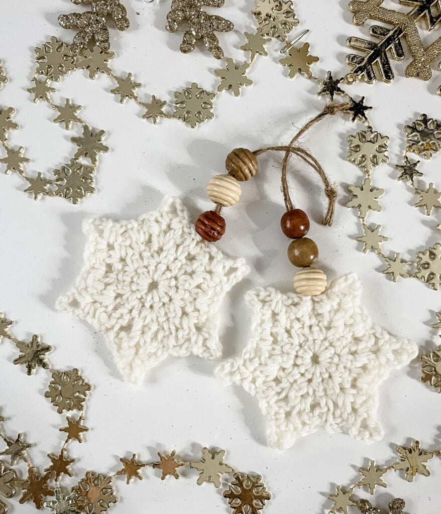Two crocheted white snowflake ornaments with wooden beads and twine—classic beaded snowflake ornaments—are surrounded by gold and glittery snowflake confetti on a white surface.