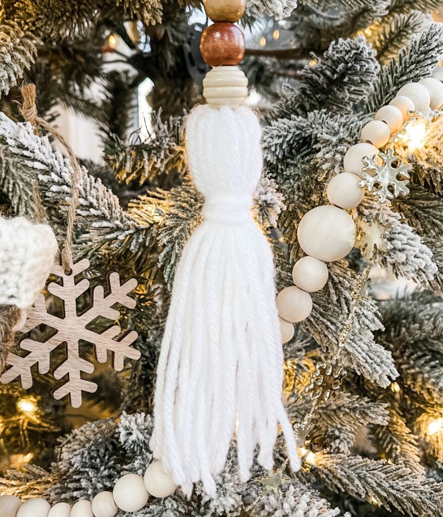Boho Beaded Tassel Ornament featuring white yarn and wooden beads hangs on a snow-dusted Christmas tree, accented with a wooden snowflake and glowing string lights.