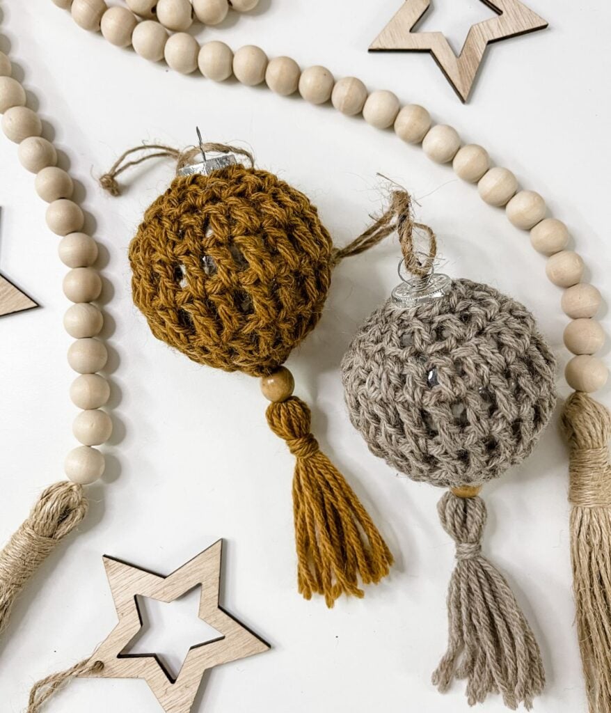 Two Boho Christmas Ball ornaments wrapped in crocheted covers, one mustard and one beige with tassels, are nestled among wooden beads and star-shaped wooden decorations.