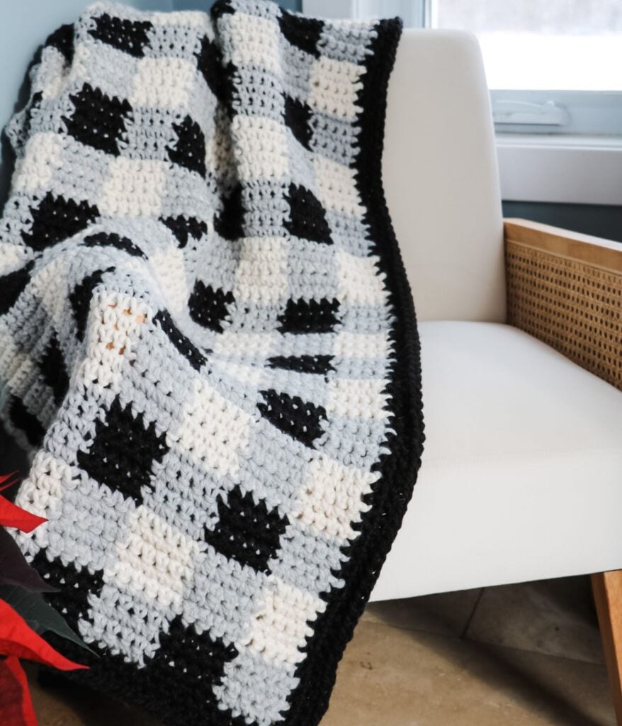 Chunky Buffalo Plaid Crochet Throw in black, white, and gray checkered pattern draped over a white chair next to a window.