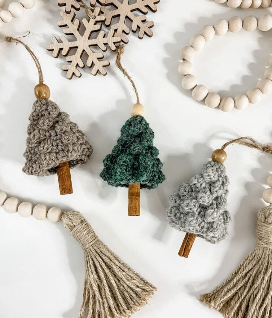 Three crocheted Cinnamon Bobble Tree ornaments with wooden bead tops and cinnamon stick trunks are arranged on a white surface with wooden beads, wood snowflakes, and twine tassels.