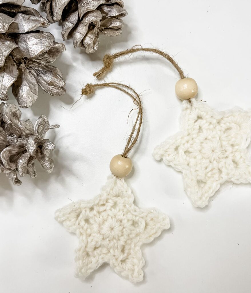 Two Crochet Star Ornaments with wooden beads and twine loops are placed next to silver-painted pine cones on a white surface.