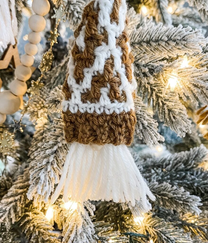 Close-up of a crocheted brown and white Mini Mosaic Gnome ornament with fringe, hanging on a snow-frosted Christmas tree with warm white lights.