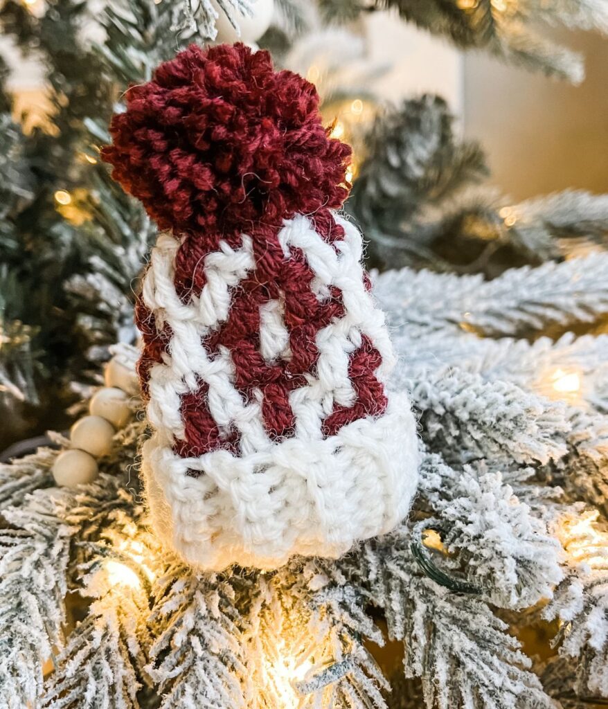 A small knitted Mini Mosaic Hat ornament, shaped like a winter hat with a red pom-pom, hangs on a snow-covered Christmas tree branch adorned with twinkling lights.