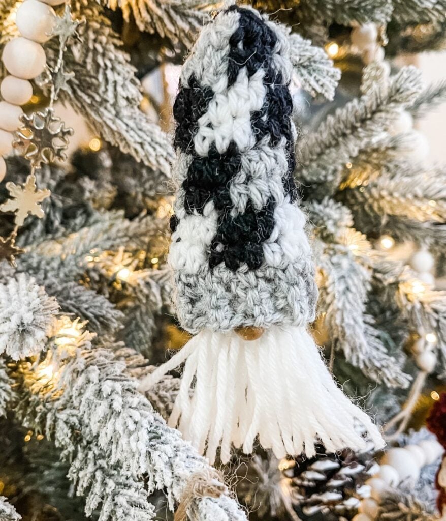 A Mini Plaid Gnome ornament with a black and white hat and white yarn beard hangs on a snowy Christmas tree branch.