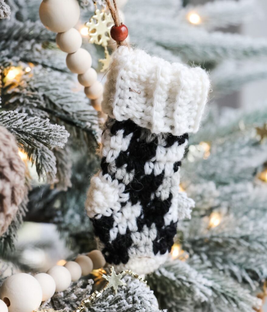 A black and white knitted mitten ornament and a mini plaid stocking hang on a snow-dusted Christmas tree decorated with wooden bead garland and string lights.