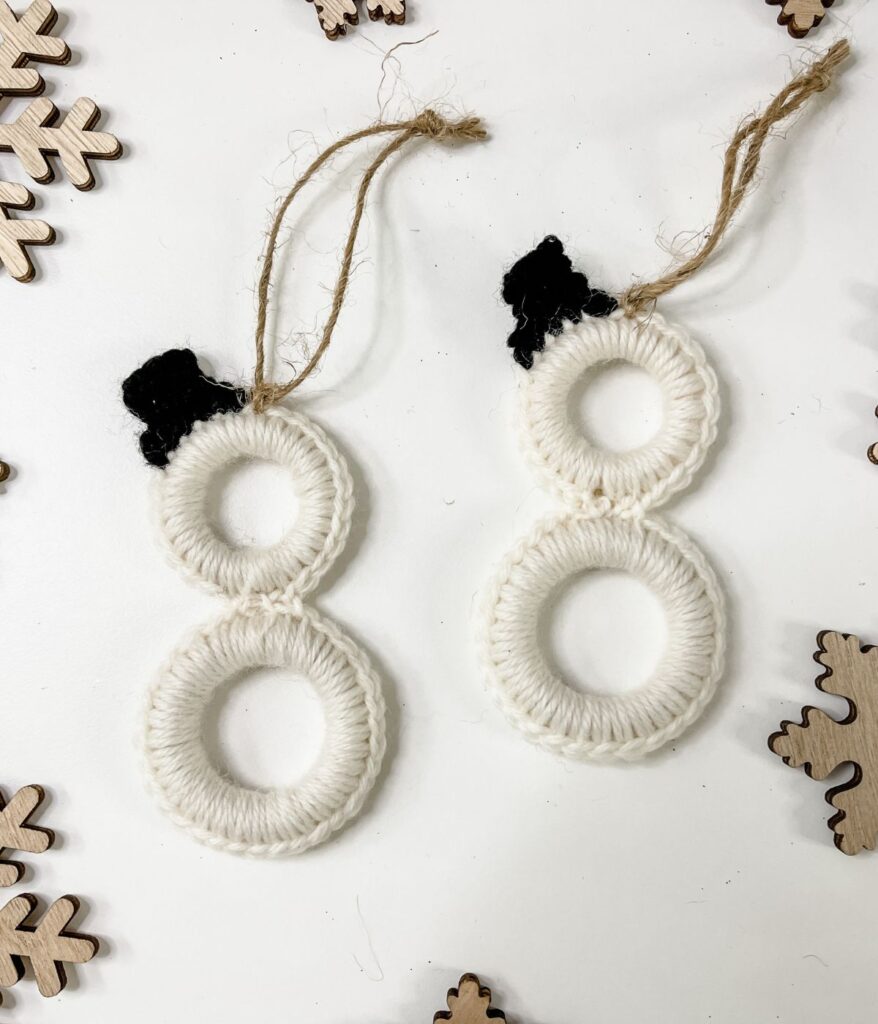 Two handmade Mini Ring Snowman ornaments crafted from white yarn wrapped around rings, with black yarn hats and twine loops for hanging, are displayed on a white surface surrounded by wooden snowflakes.