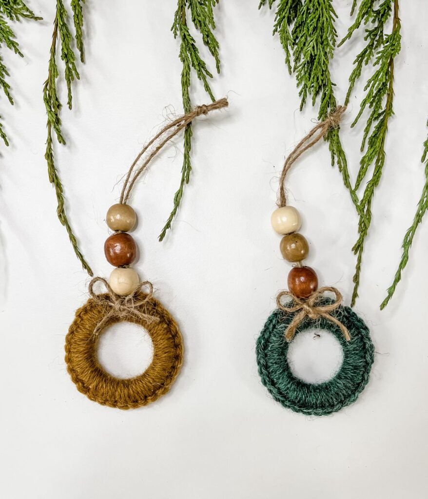 Two handmade Mini Ring Wreath ornaments with yarn-wrapped rings, wooden beads, and twine bows are displayed on a white surface with green pine branches above them.