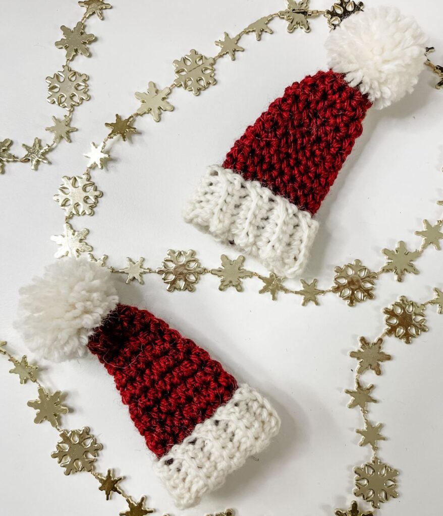 Two mini Santa hats, crocheted with white pom-poms and trim, rest on a white surface adorned with gold snowflake garlands.