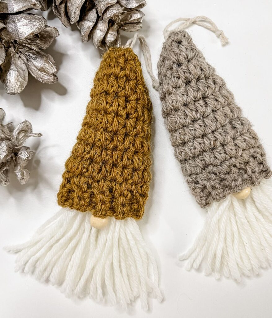 Two crocheted Mini Simple Gnome Ornaments with white yarn beards and wooden noses, one brown and one gray, rest beside painted pinecones on a white surface.