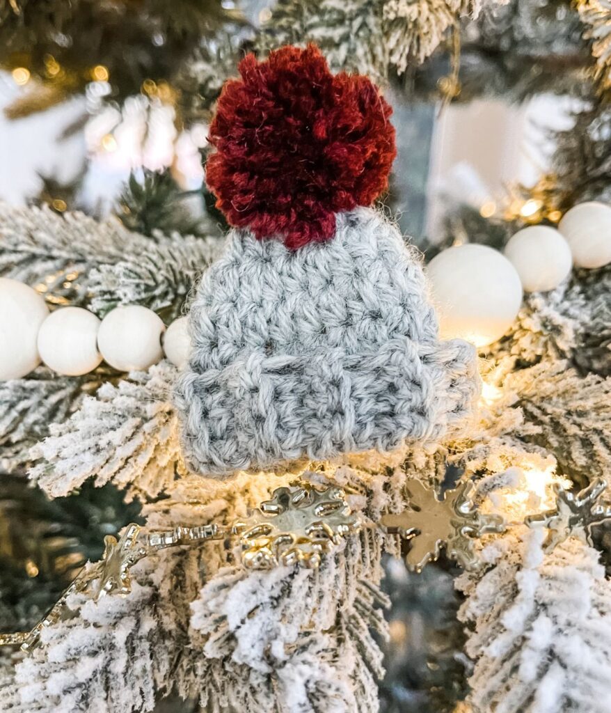 A Mini Simple Hat Ornament, shaped like a winter hat with a red pom-pom, sits on a snowy Christmas tree branch beside white bead garland and glowing string lights.