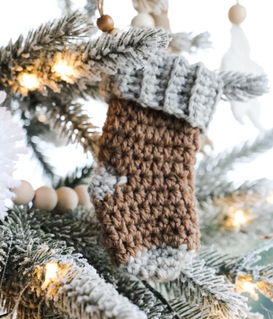 A crocheted mitten ornament in brown and gray hangs beside a Mini Simple Stocking on a snow-frosted Christmas tree branch adorned with lights and wooden bead garland.