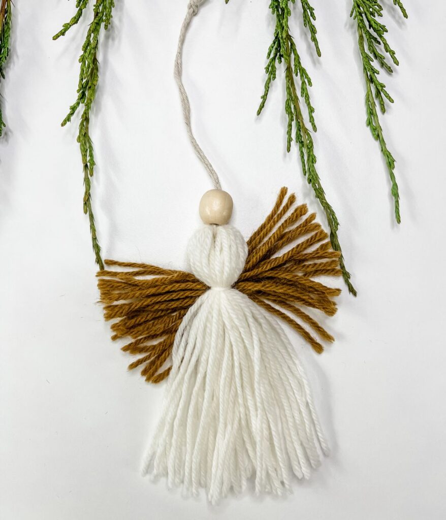 Mini Tassel Angel Ornament featuring a white yarn body, brown wings, a round wooden bead for the head, and a loop for hanging, displayed against a white background with greenery.
