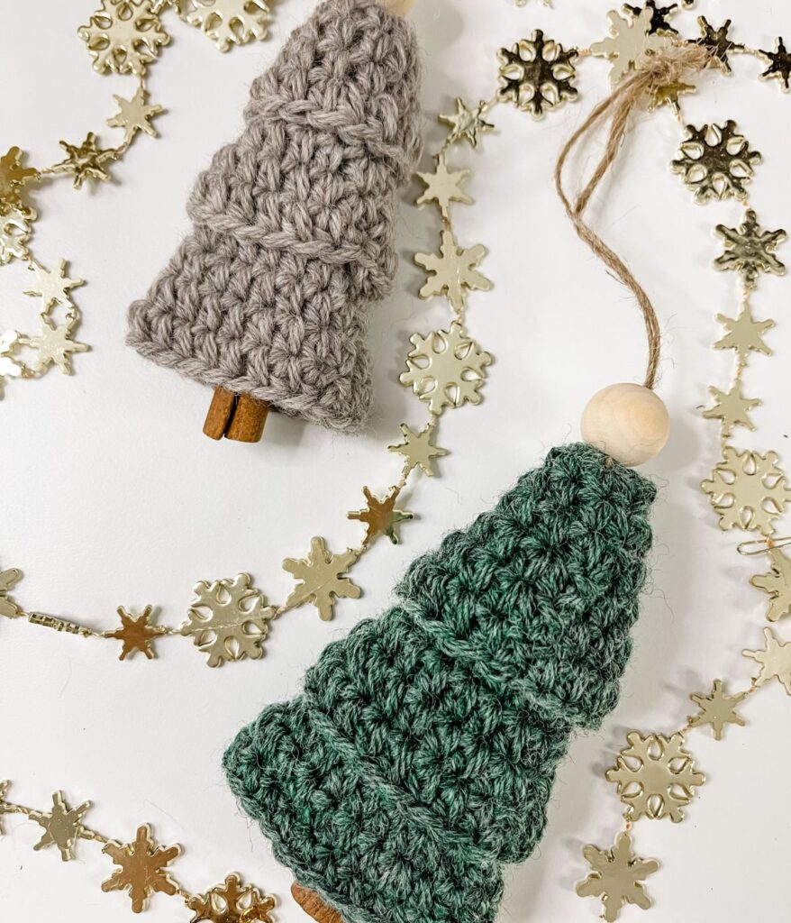 Two crocheted Mini Tiered Tree Ornaments with wooden bead tops and cinnamon stick trunks, surrounded by gold snowflake decorations on a white surface.