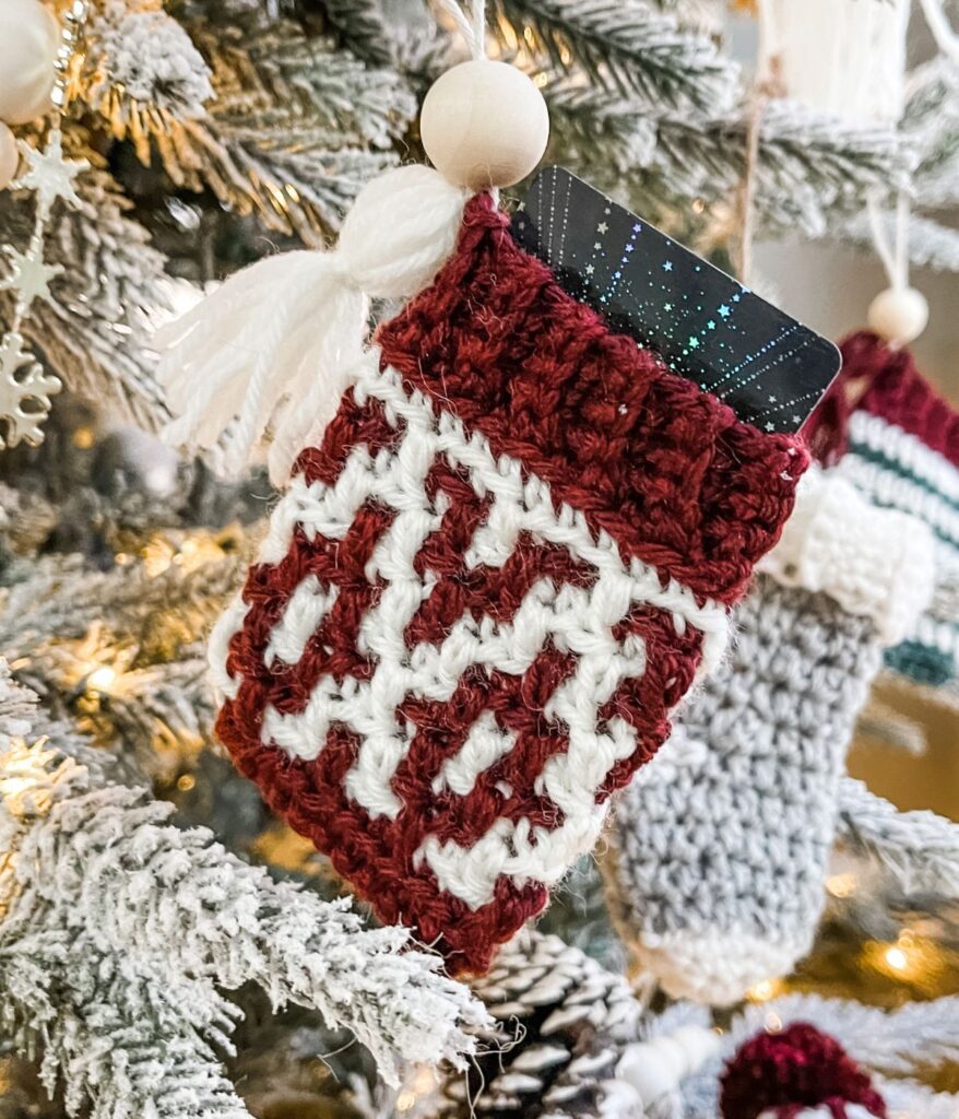 A small red and white knitted Mosaic Gift Card Cozy ornament hangs on a snowy Christmas tree branch, holding a black gift card.