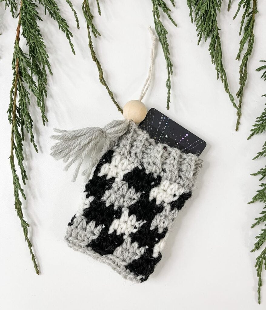 A Plaid Gift Card Cozy, crocheted in black, white, and gray with a tassel and wooden bead, holds a gift card on a white surface surrounded by green foliage.