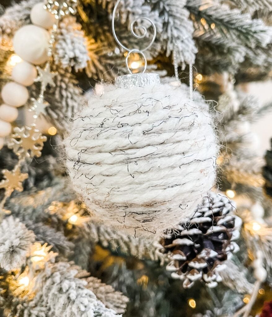 A Scrappy Christmas Ball ornament, wrapped in white yarn, hangs on a Christmas tree with frosted branches and a pine cone, surrounded by lights and decorations.