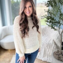 A woman with long, wavy hair wearing a cream-colored 1 Piece V Stitch Pullover and distressed jeans smiles in a bright, plant-decorated room.