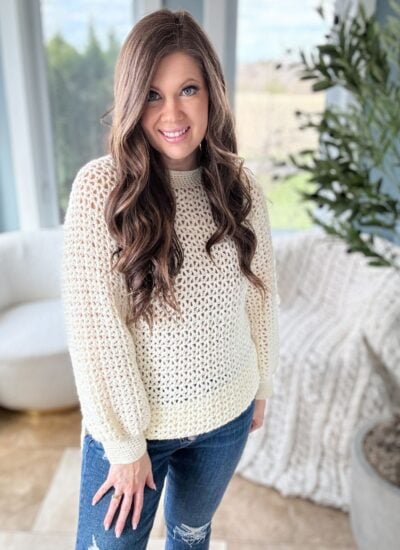 A woman with long, wavy hair wearing a cream-colored 1 Piece V Stitch Pullover and distressed jeans smiles in a bright, plant-decorated room.